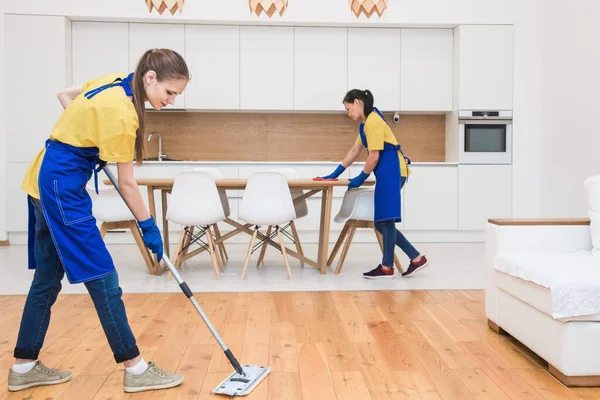 Professionele schoonmaak. Twee vrouwen in werkkleding, in schorten, verdelen de schoonmaak van de keuken van een privéhuis, huisje. koelkast wassen, kraan, gootsteen. Wasvloer. — Stockfoto