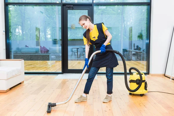 Servicio de limpieza profesional. mujer en uniforme y guantes hace la limpieza en una cabaña. el trabajador aspira el suelo con equipo profesional — Foto de Stock