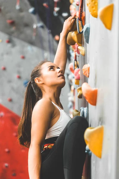 Deportiva escaladora ascendiendo sobre roca escarpada, escalando en pared artificial en interiores. Concepto de deportes extremos y bouldering — Foto de Stock