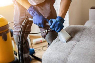 Temizlik servisi. Eldivenli ve elektrikli süpürgeli adam. Profesyonel ekipmanlı temiz kanepe.