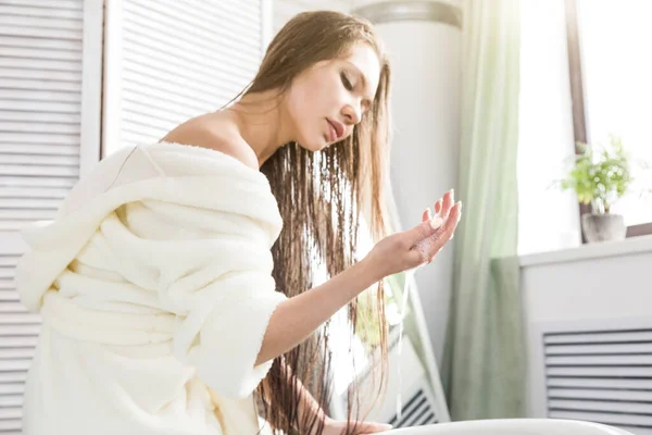 Привлекательная молодая девушка в белом халате наносит крем на кожу, сидя в салоне ванной комнаты и делая утренние процедуры. Уход за кожей и телом — стоковое фото