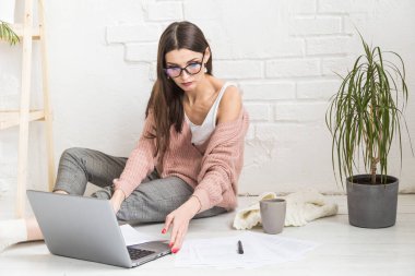 Genç ve mutlu bir kadın parlak bir apartman dairesinde ya da ofis içinde yerde oturuyor ve bir dizüstü bilgisayarda çalışıyor, iş yerinde serbest çalışan bir kız, uzaktan öğrenme öğrencileri, evrak işleri ve çevrimiçi öğrenim kavramı