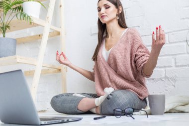 Genç ve mutlu bir kadın, parlak bir dairede yoga dairesinde yerde oturuyor ve dizüstü bilgisayarda çalışıyor. Serbest çalışan bir kız meditasyon yaparken iş strese giriyor.