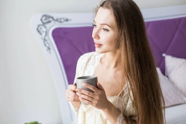 Menina atraente em roupa de noite senta-se na borda da cama junto à janela com uma xícara de café — Fotografia de Stock