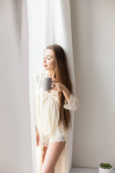 魅力的な若いです女の子で夜服に立っていますウィンドウによってコーヒーのマグカップ — ストック写真