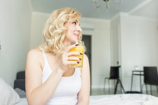 Aantrekkelijke blonde vrouw zit aan de rand van het bed thuis en houdt een kous met sinaasappelsap in haar hand. Ochtendstemming en kracht — Stockfoto