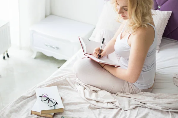 Mulher loira atraente senta-se na beira da cama em casa e escreve em um caderno — Fotografia de Stock