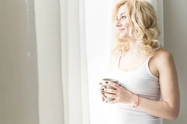 Portret van een aantrekkelijke blonde vrouw die bij het raam staat met een kop koffie of thee — Stockfoto