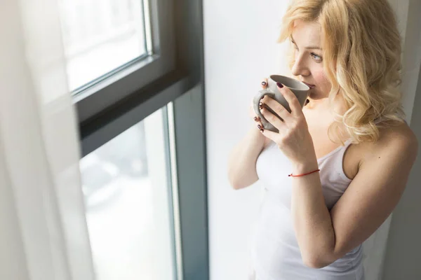 Aantrekkelijke blonde vrouw staat bij het raam met een kopje koffie of thee — Stockfoto