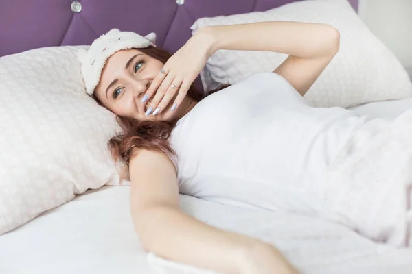 Een aantrekkelijke jonge brunette meisje vrouw ontwaakt en nipt terwijl geeuwen in haar bed in een slaapmasker — Stockfoto
