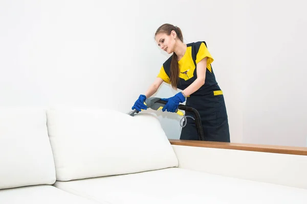 Professioneller Reinigungsservice. Zwei Frauen in Arbeitsuniform, in Schürzen. Zwei Frauen in Arbeitsuniform, in Schürzen, teilen sich die Reinigung von Privathaus, Ferienhaus. Waschen, Staubsaugen auf dem Sofa — Stockfoto