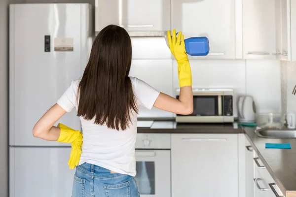 Uma jovem de uma empresa de limpeza profissional limpa em casa. Um homem lava a cozinha com luvas amarelas com material de limpeza. . — Fotografia de Stock