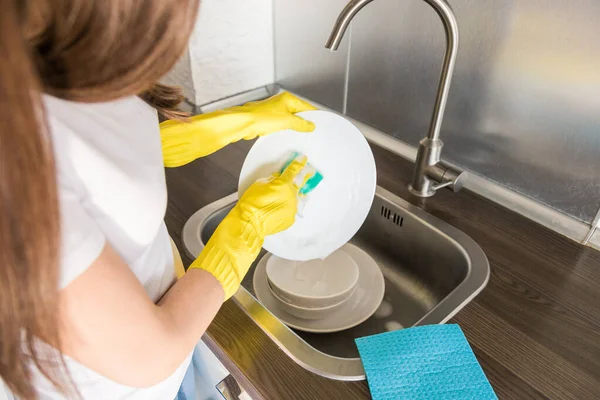 Молодая женщина в жёлтых перчатках моет посуду с губкой в раковине. Услуги по уборке дома . — стоковое фото