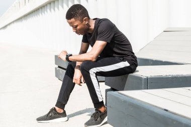 Atletik genç Afroamerikalı adam gezinti güvertesinde koşuyor. Siyah erkek koşucu dışarıda koşuyor. Sağlıklı yaşam tarzı kavramı. Adam akıllı saatle nabzına bakıyor.