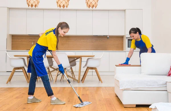 Servicio de limpieza profesional. Dos mujeres en uniforme de trabajo, en delantales, dividen la limpieza de la cocina de una casa privada, casa de campo. lavar el refrigerador, grifo, fregadero. Lavar el suelo. — Foto de Stock