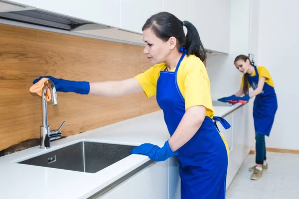Professioneller Reinigungsservice. Zwei Frauen in Arbeitsuniform, in Schürzen, teilen sich die Reinigung der Küche eines Privathauses, eines Ferienhauses. Kühlschrank waschen, Wasserhahn abstellen, Spüle. Waschboden. — Stockfoto