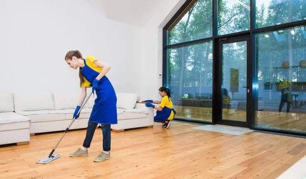 Servicio de limpieza profesional. Dos mujeres en uniforme de trabajo, en delantales, dividen la limpieza de la cocina de una casa privada, casa de campo. Suelo de lavado —  Fotos de Stock