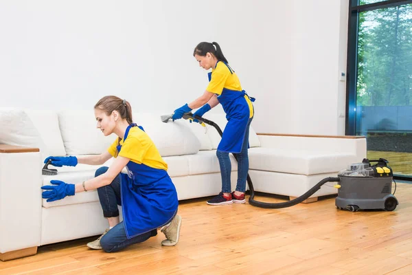 Profesyonel temizlik servisi. Önlüklü, işçi üniformalı iki kadın. Önlüklü, işçi üniformalı iki kadın, özel evin temizliğini bölüşüyorlar. Yıkama, süpürme, kanepeyi temizleme. — Stok fotoğraf