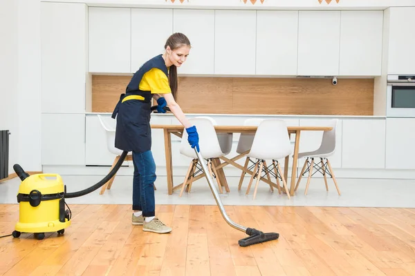 Servicio de limpieza profesional. mujer en uniforme y guantes hace la limpieza en una cabaña. el trabajador aspira el suelo con equipo profesional — Foto de Stock