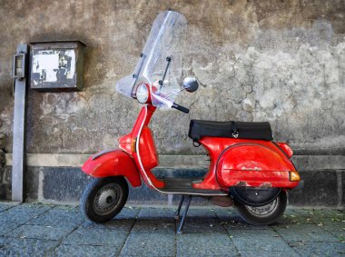 Kaldırımda beton bir duvarın yanına park edilmiş klasik kırmızı scooter. Araçta paslı çizikler ve göçükler var..