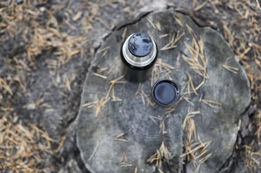 Siyah termos şişesi ve bir fincan çay devrilmiş çam ağacı iğneleriyle çevrili bir kütüğün üzerinde duruyor. Sonbahar parkında piknik.