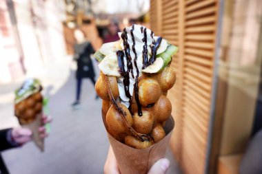 Meşgul bir caddenin bulanık arka planında waffle, meyve ve çikolatalı dondurma külahını tutan bir kadın. Seçici odak