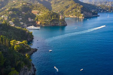 Yatlar ve tekneler İtalya 'nın Portofino kentindeki dağlık arka planda Ligurian Denizi' nde yol alıyorlar. Üzerinde geleneksel İtalyan evleri olan kayalar ve tepeler.