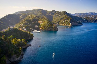 Yatlar ve tekneler İtalya 'nın Portofino kentindeki dağlık arka planda Ligurian Denizi' nde yol alıyorlar. Üzerinde geleneksel İtalyan evleri olan kayalar ve tepeler.