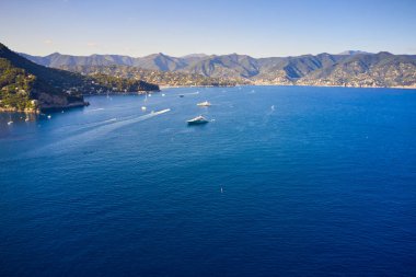 Yatlar ve tekneler İtalya 'nın Portofino kentindeki dağlık arka planda Ligurian Denizi' nde yol alıyorlar. Üzerinde geleneksel İtalyan evleri olan kayalar ve tepeler.