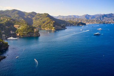 Yatlar ve tekneler İtalya 'nın Portofino kentindeki dağlık arka planda Ligurian Denizi' nde yol alıyorlar. Üzerinde geleneksel İtalyan evleri olan kayalar ve tepeler.