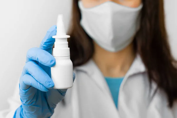 A woman using nose spray for flu treat or running nose holds medicine in the hand. Doctor in protective mask and blue gloves shows nose spray for illness treatment
