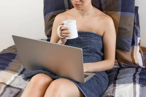 Work from home during quarantine. Young woman in a blue towel is typing on her laptop with a white cup in her hands. Relaxing by watching movies after bath