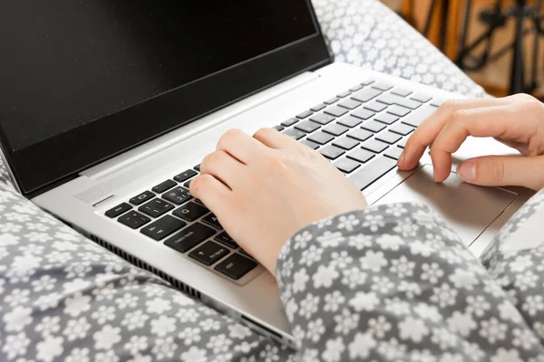 Front view on a screen of a laptop on the knees of a woman in a grey flowered nightwear. Home office concept. Freelancer works in a bed during outbreak of coronavirus.