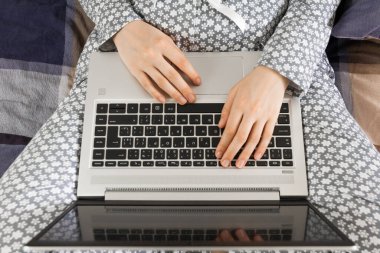 Yakın plan kadın elleri dizüstü bilgisayarın klavyesinde. Çiçekli gri gecelikli kadın internette bir yatakta çalışıyor. Freelancer bir deftere metin yazıyor.