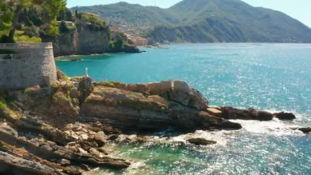 Survolez la côte rocheuse avec une statue blanche de Jésus au sommet et des montagnes à l'arrière-plan. Une plage avec un mur de forteresse est baignée par de l'eau turquoise bleue verte. D'énormes pierres sous l'eau
