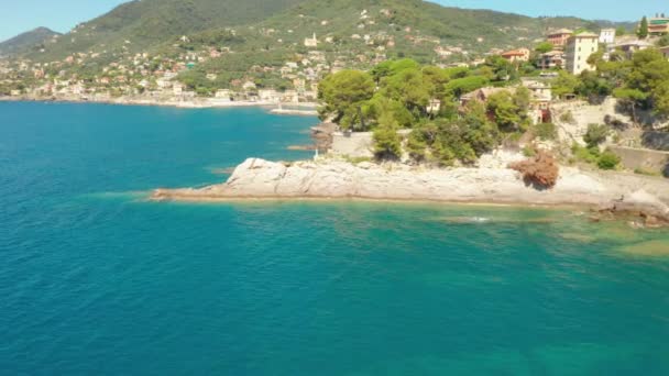 Vlieg over de rotsachtige kust met een wit standbeeld van Jezus op de top en bergen op de achtergrond. Traditionele Italiaanse kleurrijke huizen op de heuvel. Enorme stenen onder water Camogli, Italië. — Stockvideo