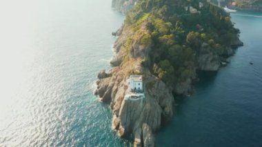 İtalya 'nın Portofino kentindeki yeşil ağaçlı ve villalı kayalık tepenin üzerinden geçen deniz fenerinden görüntüler. Ligurian Denizi bir tepenin etrafında turkuaz suyla çevrilidir..