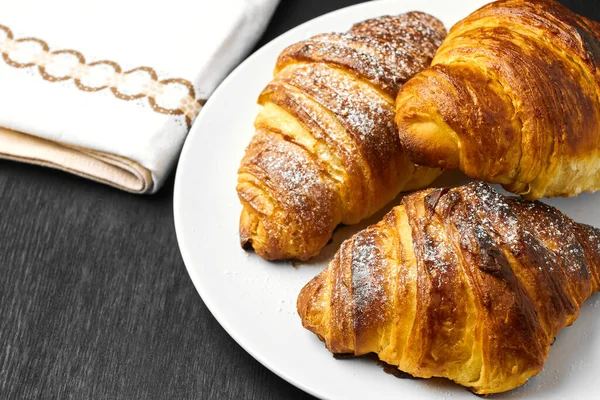 Croissants lisos recém-assados em uma placa branca perto de serviette em um fundo de madeira preto. Pastelaria francesa caseira . — Fotografia de Stock