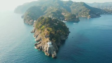 Ligurian denizindeki kayalık bir tepenin üzerinde duran deniz fenerinin görüntülerinde mavi turkuaz su ve arka planda küçük bir motorlu tekne, Portofino, İtalya. 