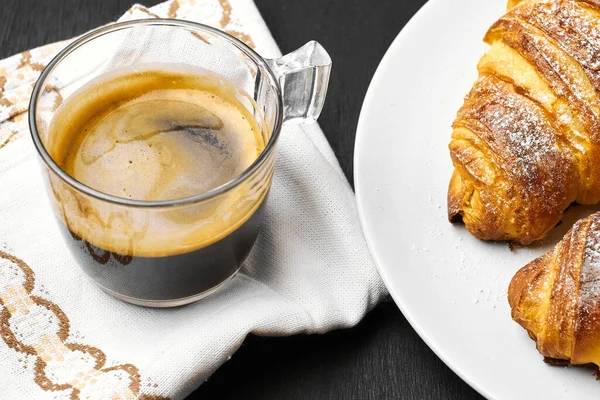 Una tazza di espresso su un tovagliolo di cotone, croissant burrosi su fondo nero. Fatto in casa francese cottura — Foto Stock