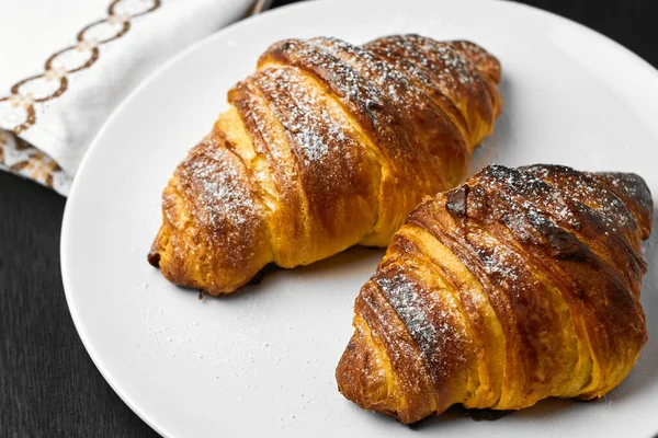Croissants lisos recém-assados em uma placa branca perto de serviette em um fundo de madeira preto. Pastelaria francesa caseira . — Fotografia de Stock