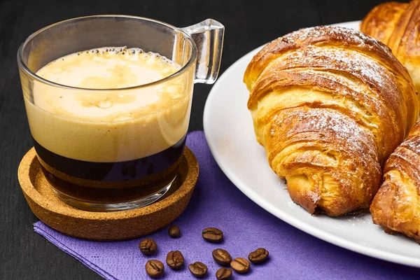 Feche uma xícara de café expresso e croissants frescos em um serviette roxo com grãos de café. Conceito de café da manhã em um fundo preto — Fotografia de Stock