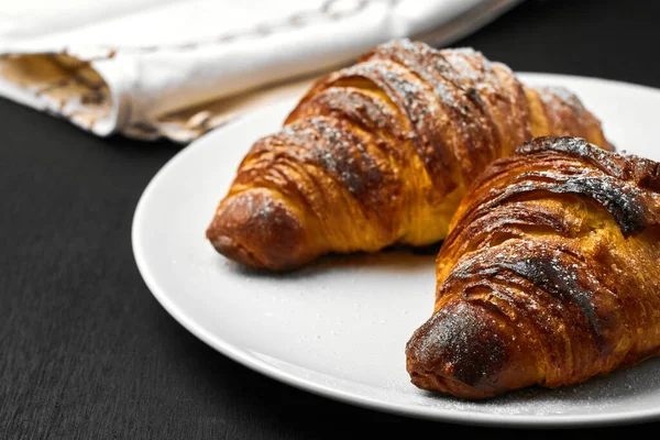 Croissants lisos recém-assados em uma placa branca perto de serviette em um fundo de madeira preto. Pastelaria francesa caseira . — Fotografia de Stock