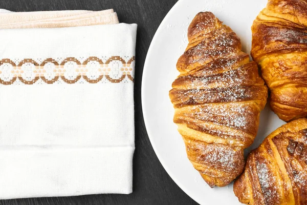 Croissants lisos recém-assados em uma placa branca perto de serviette em um fundo de madeira preto. Pastelaria francesa caseira com espaço de cópia — Fotografia de Stock
