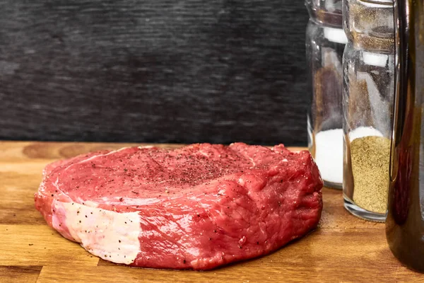 Peça crua fresca de uma carne de vaca ao lado de sal e pimentão em garrafas de vidro em um contexto preto. Conceito de recepção . — Fotografia de Stock