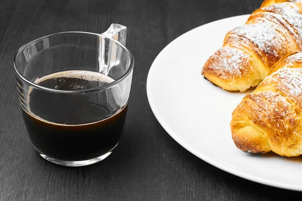 Feche dois croissants em uma chapa branca e uma xícara de café em um fundo preto. Conceito de pequeno-almoço italiano . — Fotografia de Stock