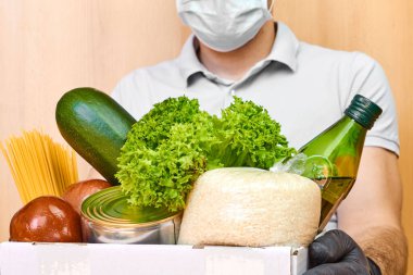 Koruyucu maskeli teslimatçı ve lastik eldivenli adam çeşitli yiyeceklerle dolu bir kutuyu tutuyor. Coronavirus karantinası sırasında yiyecek teslimatı. 