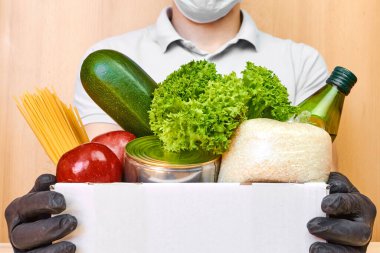 Koruyucu maskeli teslimatçı ve lastik eldivenli adam çeşitli yiyeceklerle dolu bir kutuyu tutuyor. Coronavirus karantinası sırasında yiyecek teslimatı. 