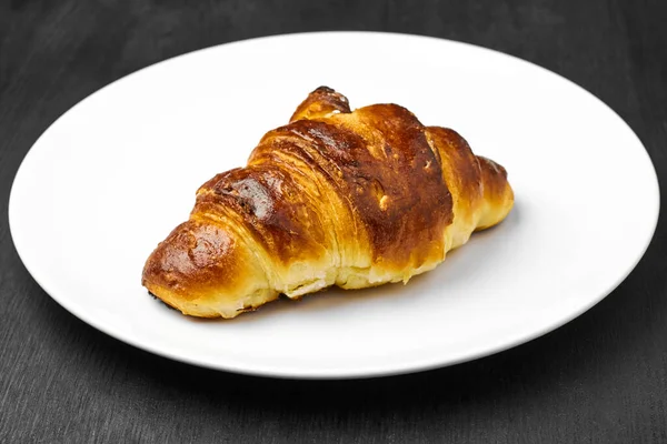 Fresh croissant with a brown crust on a white plate on a black table. — Stock Photo, Image