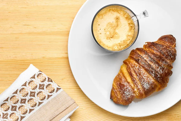 Vista superior em um café da manhã composto por uma xícara de café, um croissant em uma mesa de madeira com espaço de cópia. Serviette perto da placa branca com lanche . — Fotografia de Stock